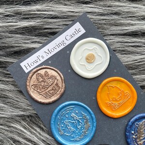 Pode incluir: Seis selos de cera com diferentes desenhos em um fundo cinza escuro. Os selos s&atilde;o redondos e apresentam uma variedade de cores, incluindo branco, dourado, cobre, azul, laranja e preto. O texto "Howl's Moving Castle" est&aacute; impresso em branco no fundo.