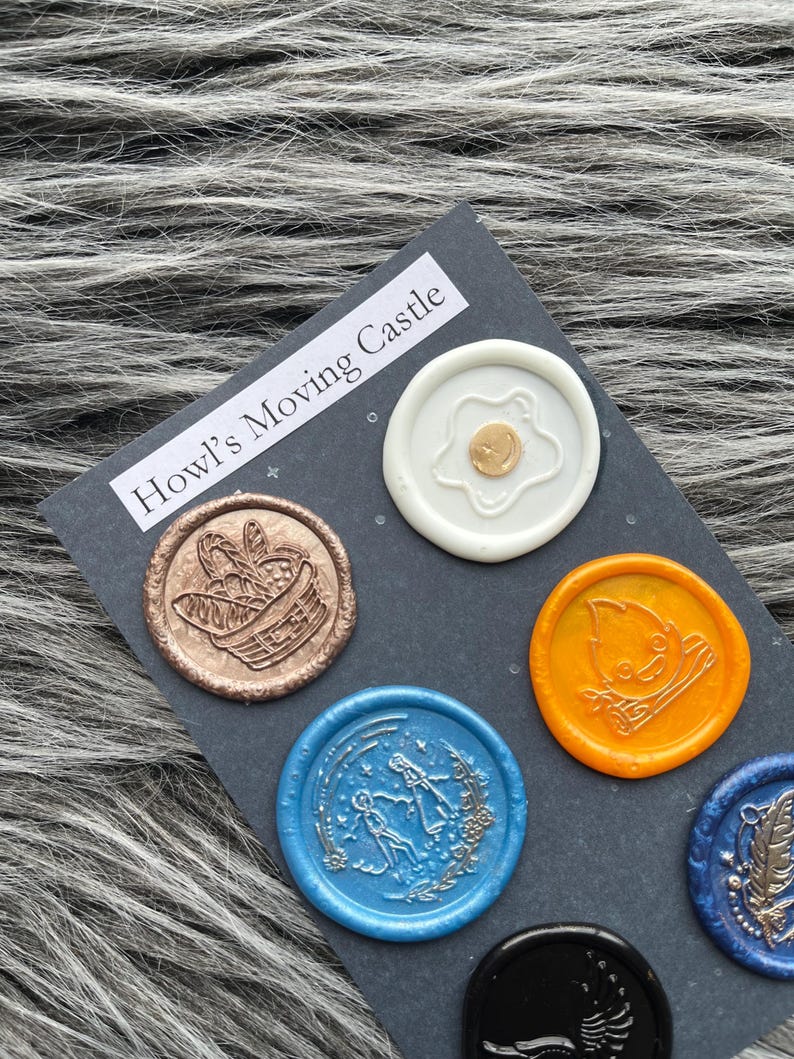 Pode incluir: Seis selos de cera com diferentes desenhos em um fundo cinza escuro. Os selos s&atilde;o redondos e apresentam uma variedade de cores, incluindo branco, dourado, cobre, azul, laranja e preto. O texto "Howl's Moving Castle" est&aacute; impresso em branco no fundo.