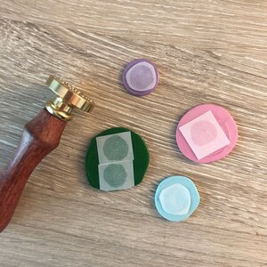 May include: A wooden-handled wax seal stamp with a gold-colored stamp head rests next to four colorful wax seals. The seals are in shades of green, pink, purple, and blue, each with a paper backing.