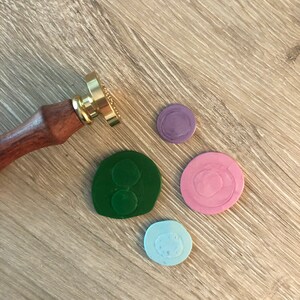 May include: A wooden-handled wax seal stamp with a gold-colored stamp head, next to four colorful wax seals in green, pink, light blue, and purple. The seals are on a light brown wooden surface.