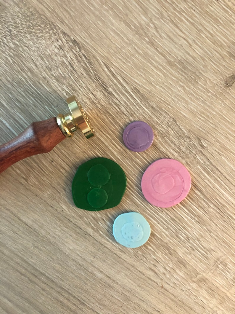 May include: A wooden-handled wax seal stamp with a gold-colored stamp head, next to four colorful wax seals in green, pink, light blue, and purple. The seals are on a light brown wooden surface.
