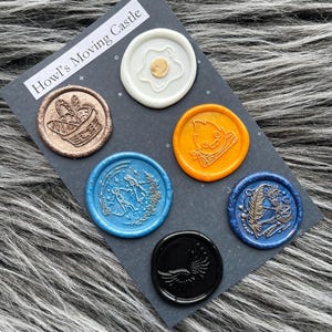 Pode incluir: Seis selos de cera com diferentes desenhos e cores. Os selos est&atilde;o em um cart&atilde;o preto com o texto "O Castelo Animado de Howl".