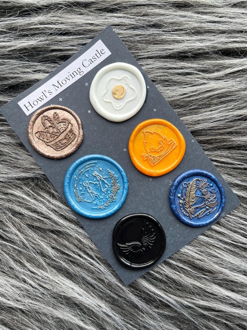Pode incluir: Seis selos de cera com diferentes desenhos e cores. Os selos est&atilde;o em um cart&atilde;o preto com o texto "O Castelo Animado de Howl".