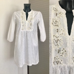 May include: White tunic dress with long sleeves and a mandarin collar. The dress features floral embroidery and sequin embellishments around the neckline. The fabric has a subtle, embossed floral pattern throughout.