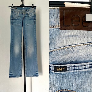 Peut inclure: Jean en denim bleu clair avec un délavage, des poches avant et une coupe évasée. Le jean est présenté sur un cintre, mettant en valeur les détails du tissu et du design. Un gros plan révèle l'étiquette de la marque "Lee".