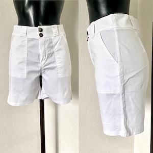 May include: White shorts with two front pockets and two brown buttons. The shorts are displayed on a mannequin. The shorts have a tailored fit and a classic design, suitable for casual wear.