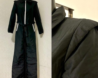 Könnte beinhalten: Ein schwarzer gesteppter Overall mit einem weißen Reißverschluss und weißen Verzierungen an den Taschen und Gürtelschlaufen. Der Overall hat eine lange, fließende Silhouette.