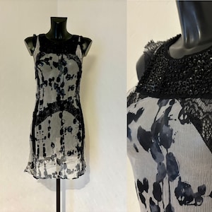 Puede incluir: Un vestido transparente blanco y negro con ribete de encaje negro y escote de cuentas. El vestido tiene un corte holgado y una silueta fluida.