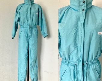 May include: A light blue ski suit with a high collar, zipper closure, and elastic waist. The suit has a logo on the left sleeve that says "Pichl".