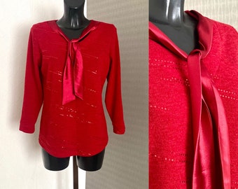 May include: A bright red long-sleeved top with a satin tie at the neckline. The top has a textured knit design with small, evenly spaced holes. The sleeves are three-quarter length. The top is displayed on a black mannequin.