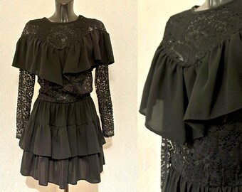 Può includere: Un abito nero in pizzo e chiffon con scollo a volant e gonna a strati. L'abito ha maniche lunghe e un corpetto aderente.