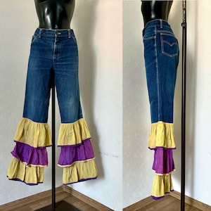 Puede incluir: Vaqueros azules con diseño de pernera acampanada. La parte inferior de los vaqueros presenta tres niveles de volantes en amarillo y morado. Los vaqueros se exhiben en un maniquí negro.