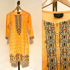 Peut inclure: Une tunique jaune à manches longues avec des broderies complexes en or, rouge et vert. La tunique a un col rond et une fermeture boutonnée à l'avant. L'étiquette indique "Made in India ALLY'S".