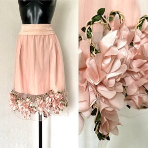Pode incluir: Uma saia de tule rosa com um design floral. A saia tem uma camada de tule com flores de tecido rosa e folhas verdes ao redor da parte inferior.