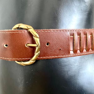 Puede incluir: Cinturón de cuero marrón con hebilla dorada. El cinturón tiene una serie de ranuras verticales recortadas a lo largo del borde superior y costuras blancas. La hebilla tiene un diseño retorcido. El cinturón se muestra en un maniquí negro.