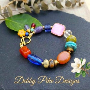 May include: A multi-colored charm bracelet featuring various beads including a pink square, blue glass, turquoise, and orange stones, accented with a yellow flower charm and a gold-toned toggle clasp. The text Debby Pike Designs is visible.