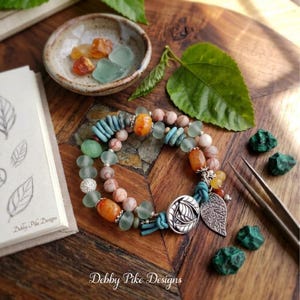 May include: A multi-strand bracelet featuring a mix of turquoise, orange, and marbled beige beads, accented with a silver-toned circular charm and a silver-toned leaf-shaped pendant, secured with a blue cord and a silver-toned button clasp.