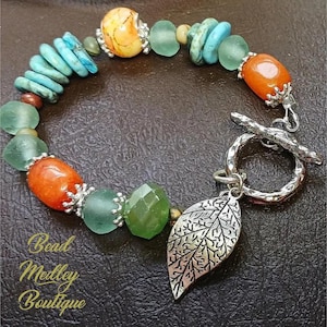 May include: A beaded bracelet featuring turquoise, orange, green, and yellow beads. The bracelet has a silver toggle clasp and a leaf charm. The text "Bead Medley Boutique" is visible.