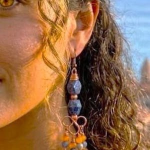 May include: A dangle earring featuring two faceted blue beads stacked vertically, connected by copper wire. Below the beads, a copper wire frame supports several smaller dangling beads in shades of orange, yellow, and light blue, creating a layered effect.