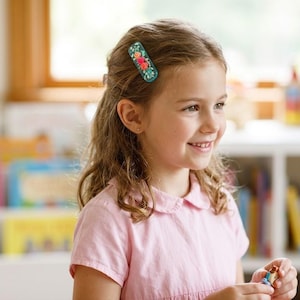 May include: A rectangular hair clip in a dark teal color featuring a floral pattern with red, pink, and white flowers and green leaves. The hair clip is secured in light brown hair, holding a section of hair back from the face.