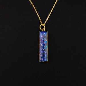 Peut inclure: Un collier en chaîne dorée avec un pendentif rectangulaire en résine bleue et violette. Le pendentif a un aspect moucheté et brillant.
