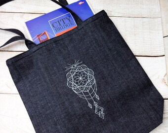 May include: A black denim tote bag with a white embroidered dreamcatcher design. The bag has a book inside with the text "CITY BRIDGE" on the cover.