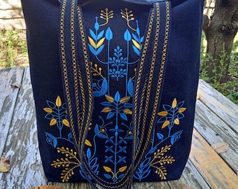 May include: A navy blue tote bag with an embroidered floral design in yellow and blue. The bag has a rectangular shape with a wide opening and two handles. The floral embroidery is centered on the front of the bag.