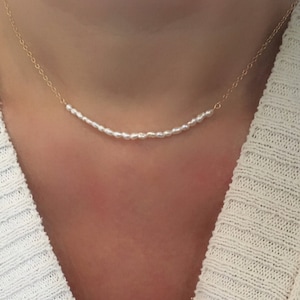 May include: A delicate gold chain necklace with a row of small white pearls. The necklace is worn by a person with a white sweater.