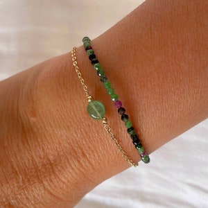 Op de afbeelding: Twee armbanden om een pols. De bovenste armband is een gouden ketting met een groene edelsteenbedel. De onderste armband is gemaakt van groene, zwarte en roze kralen.