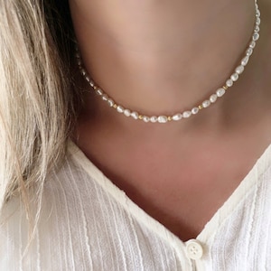 Puede incluir: Un delicado collar de perlas con pequeñas perlas blancas irregulares y cuentas doradas intercaladas. El collar se lleva sobre una blusa color crema con botones en la parte delantera.