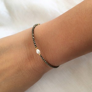 May include: A delicate bracelet featuring small, dark grey beads and a single, lustrous white pearl. Gold-coloured accents are interspersed throughout the design. The bracelet is worn on a wrist.
