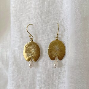 May include: A pair of gold-colored earrings. Each earring features a detailed, leaf-shaped pendant with a small, dangling pearl. The earrings are suspended from a simple hook.