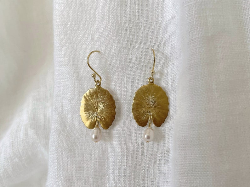 May include: A pair of gold-colored earrings. Each earring features a detailed, leaf-shaped pendant with a small, dangling pearl. The earrings are suspended from a simple hook.