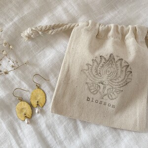 Two gold earrings with a pearl accent. The earrings are shaped like a leaf and have a delicate design. The earrings are displayed on a white fabric surface with a small white drawstring bag with the word 'blossom' printed on it.