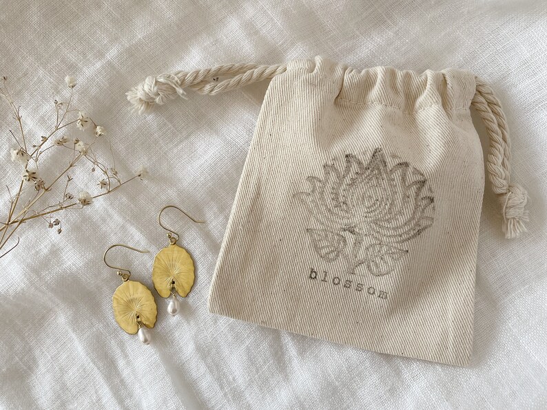 Two gold earrings with a pearl accent. The earrings are shaped like a leaf and have a delicate design. The earrings are displayed on a white fabric surface with a small white drawstring bag with the word 'blossom' printed on it.