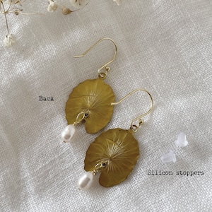 A pair of gold earrings with a leaf design and a small white pearl hanging from each. The earrings are on a white fabric background. The text 'Back' is written on the left side of the image. The text 'Silicon stoppers' is written on the right side of the image.