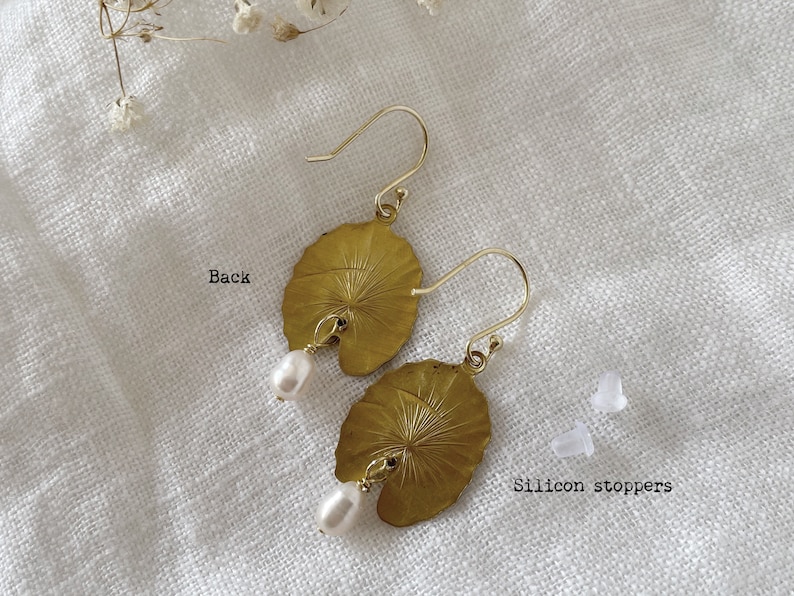 A pair of gold earrings with a leaf design and a small white pearl hanging from each. The earrings are on a white fabric background. The text 'Back' is written on the left side of the image. The text 'Silicon stoppers' is written on the right side of the image.