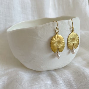 A pair of gold earrings with a pearl drop. The earrings are shaped like a leaf and have a textured surface. The earrings are displayed on a white bowl.