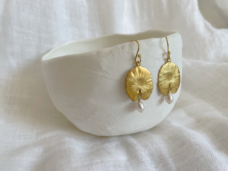 A pair of gold earrings with a pearl drop. The earrings are shaped like a leaf and have a textured surface. The earrings are displayed on a white bowl.