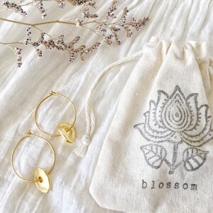 Puede incluir: Pendientes de aro dorados con un dije redondo y plano. Los pendientes se muestran sobre un fondo de tela blanca con una peque&ntilde;a rama de flores secas y una bolsa de tela blanca con cord&oacute;n con un dise&ntilde;o floral negro y la palabra "blossom" impresa en la parte delantera.