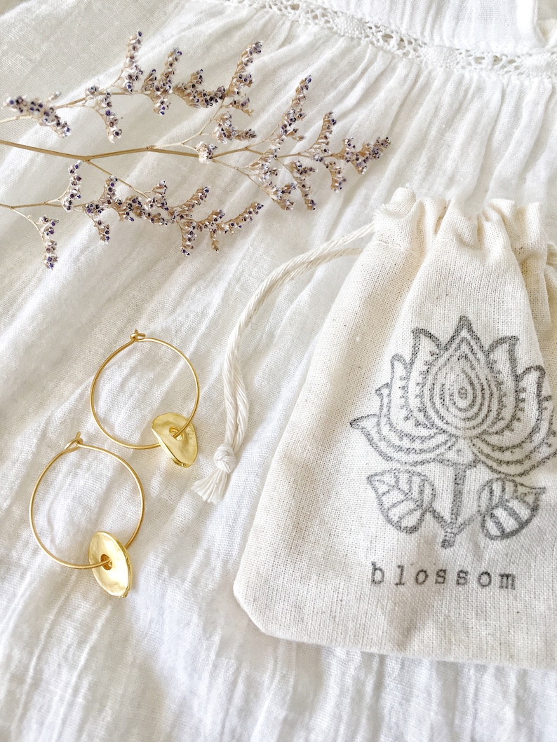 Puede incluir: Pendientes de aro dorados con un dije redondo y plano. Los pendientes se muestran sobre un fondo de tela blanca con una peque&ntilde;a rama de flores secas y una bolsa de tela blanca con cord&oacute;n con un dise&ntilde;o floral negro y la palabra "blossom" impresa en la parte delantera.