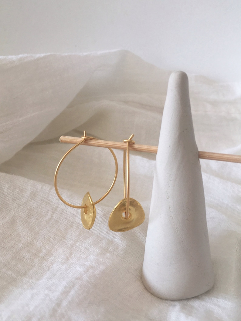 Puede incluir: Pendientes de aro dorados con un colgante de disco asim&eacute;trico texturizado. Los pendientes se muestran en un soporte de cer&aacute;mica blanca.