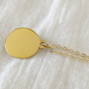 A gold pendant necklace with a round, smooth, gold disc charm. The chain is a delicate, double-strand chain.