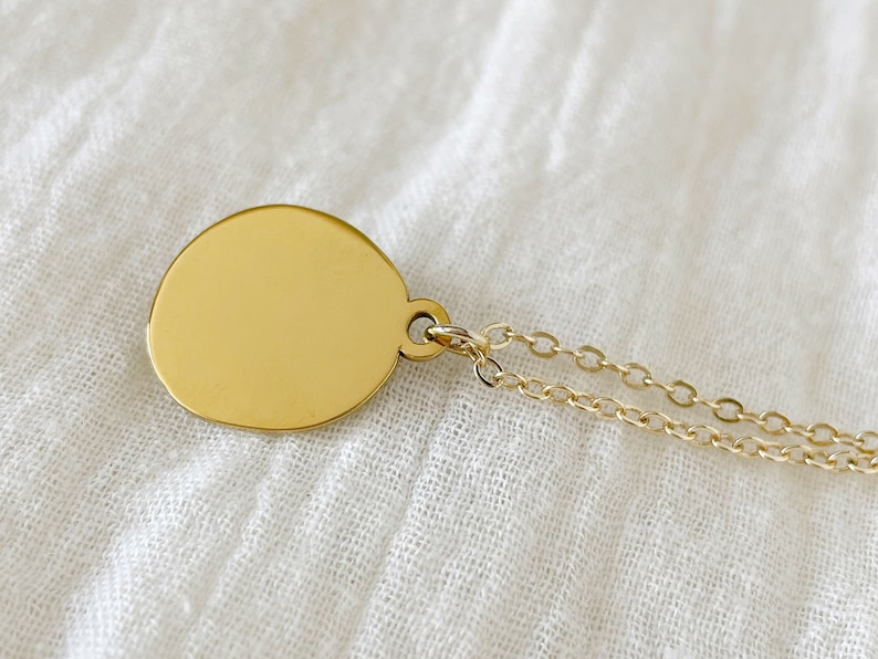 A gold pendant necklace with a round, smooth, gold disc charm. The chain is a delicate, double-strand chain.