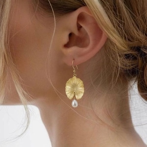 May include: Gold-colored earrings featuring a lily pad design with a dangling white pearl. The earrings are worn on a person's ear, with the lily pad shape detailed with textured lines.