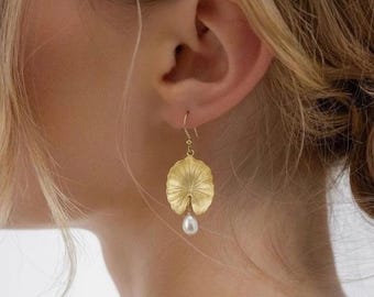 May include: Gold-colored earrings featuring a lily pad design with a dangling white pearl. The earrings are worn on a person's ear, with the lily pad shape detailed with textured lines.