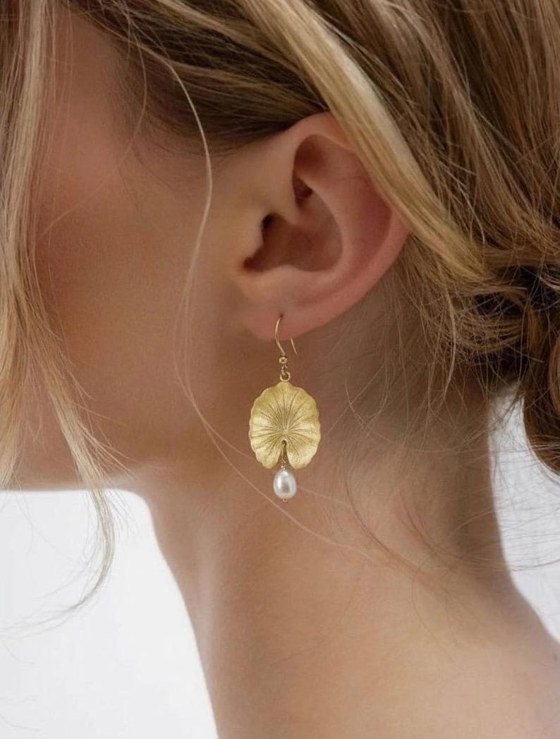 May include: Gold-colored earrings featuring a lily pad design with a dangling white pearl. The earrings are worn on a person's ear, with the lily pad shape detailed with textured lines.