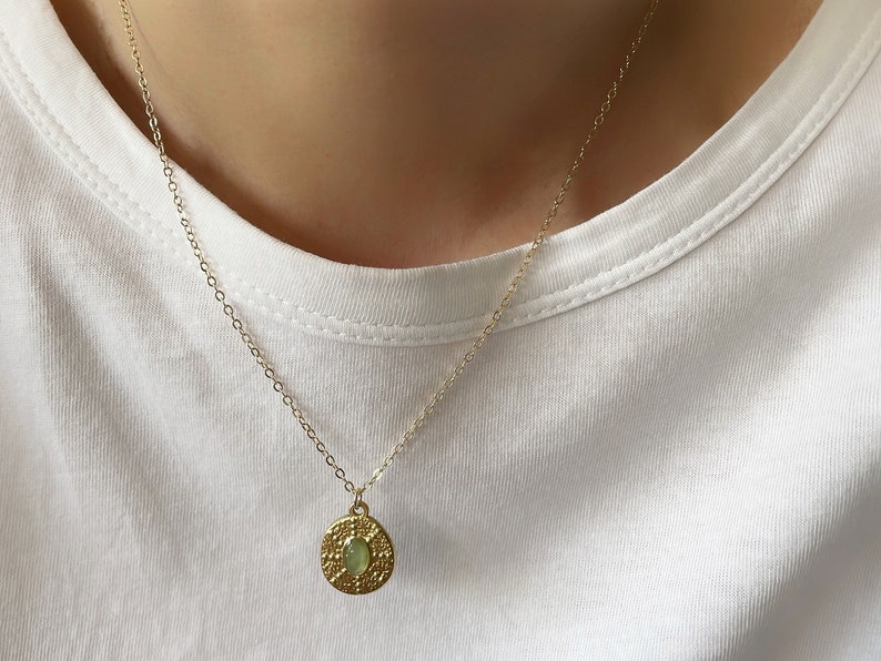 A dainty gold chain necklace with a round pendant featuring an oval shaped green gemstone.