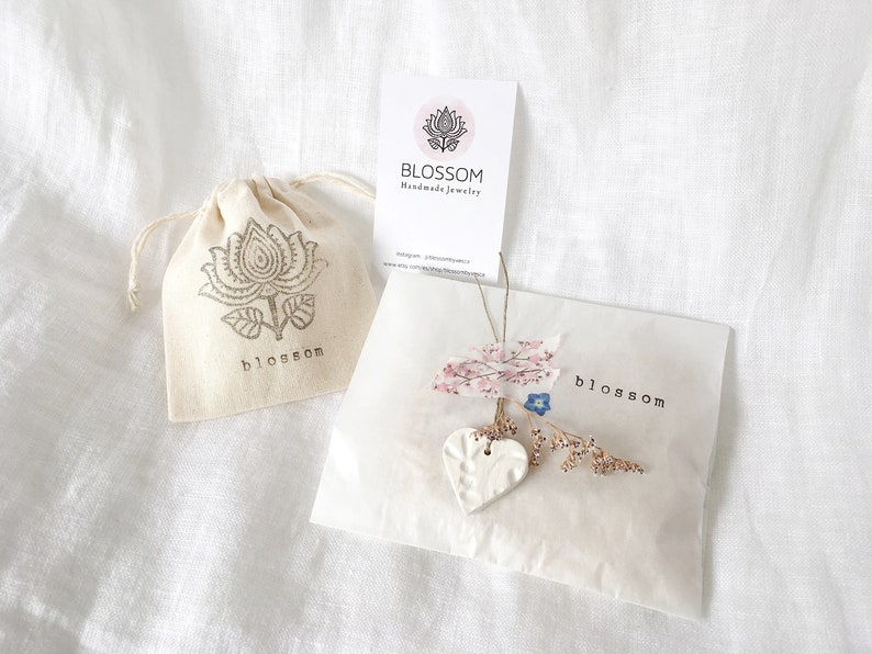 May include: A small, cream-colored drawstring bag and a white paper package with the word "blossom" printed on them. The bag has a floral design and the word "blossom" printed on it. The package is decorated with dried flowers and a ceramic heart.