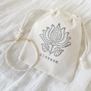 May include: A delicate pearl bracelet with a gold clasp, displayed on a white fabric pouch with a black floral design and the word "blossom" printed on it.